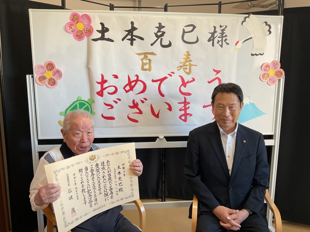 百寿祝いの行事。高齢入居者と職員が記念撮影で祝いの証書を持つ